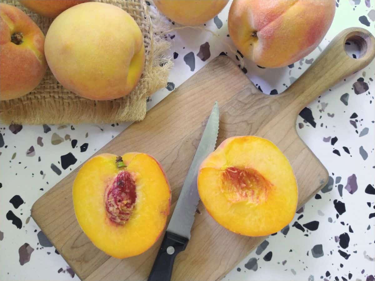 A cut open freestone peach sitting on a cutting board with a knife along with some other whole peaches. .
