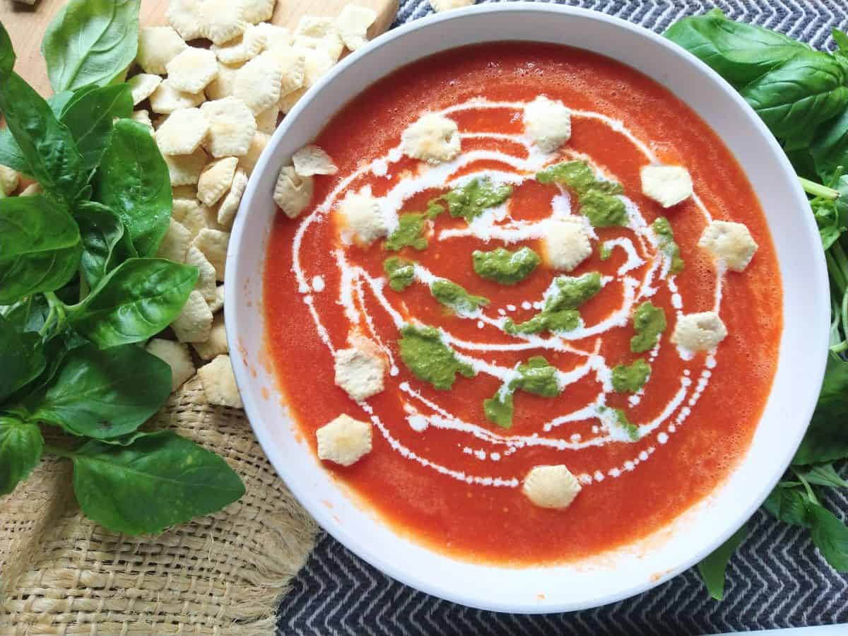 A white bowl filled with tomato soup that is topped with heavy cream, pesto, and oyster crackers. The soup is surrounded by fresh basil.