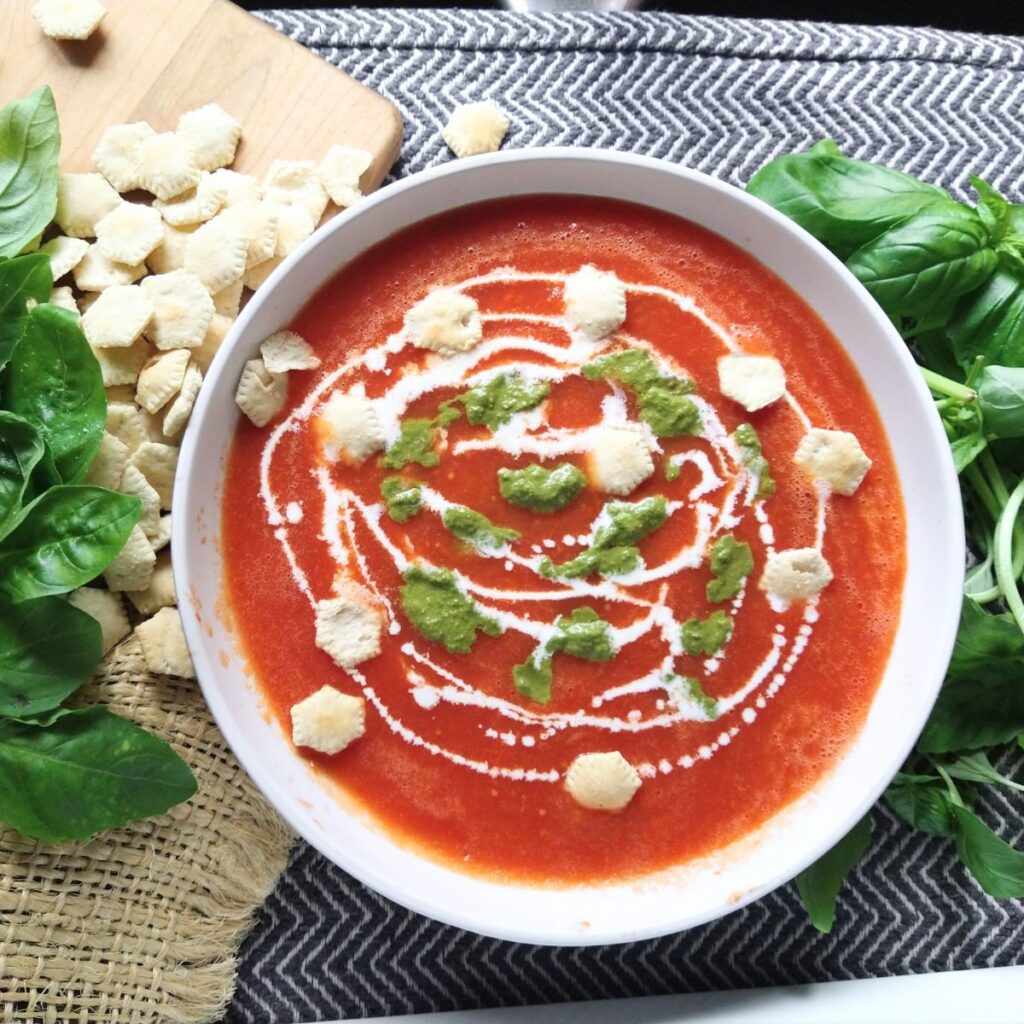 A bowl of tomato soup that is topped with cream, pesto, and oyster crackers.