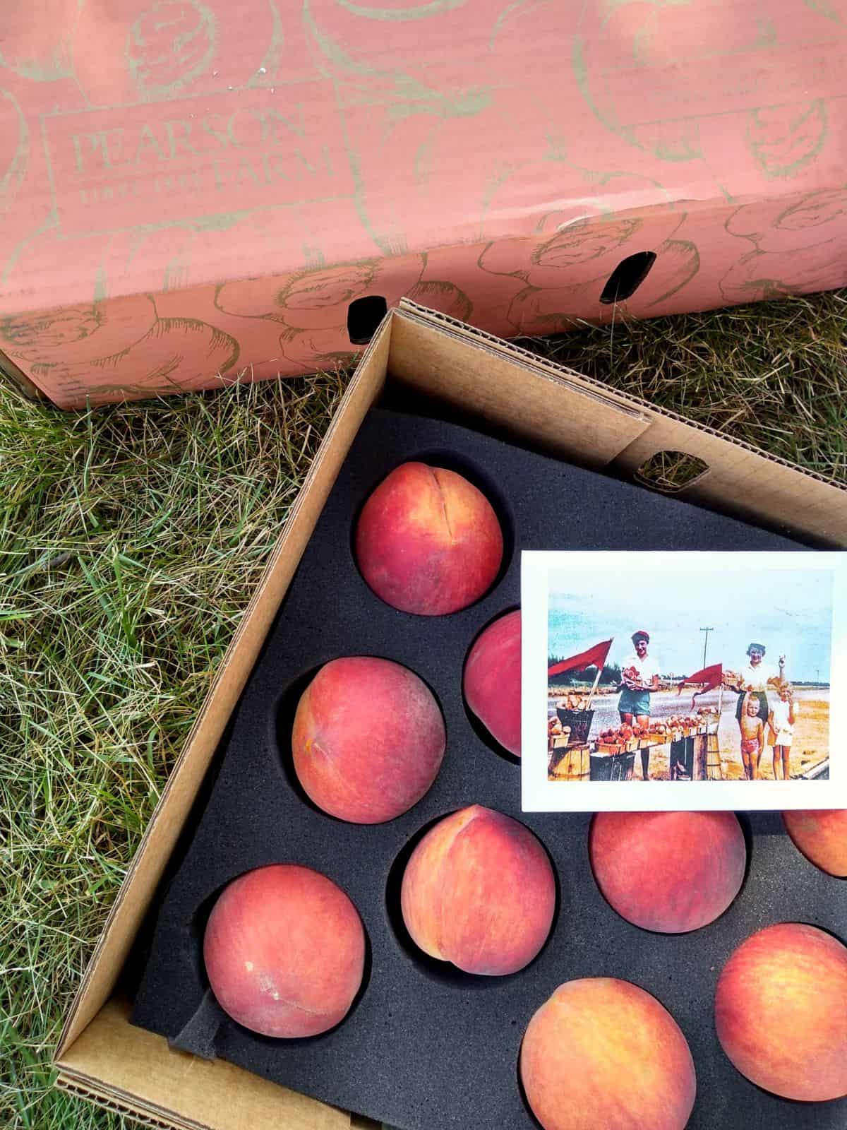 An opened box of peaches sitting on the grass. There is an old photo on top of the peaches.