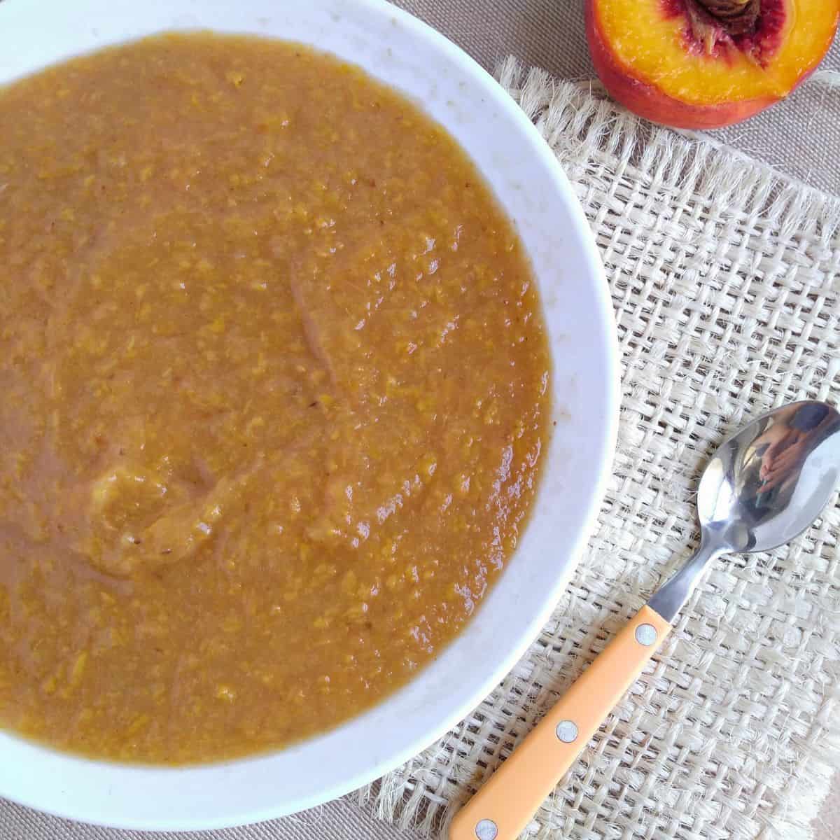 A white bowl with peach applesauce in it on top of a piece of burlap with a spoon with a peach colored handle next to it and a halved peach.