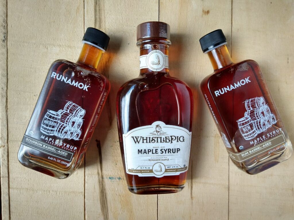 Runamok barrel aged syrups on a wood board. From left to right - Bourbon barrel aged, WhistlePig Whiskey Barrel Aged, and Rum Barrel Aged.