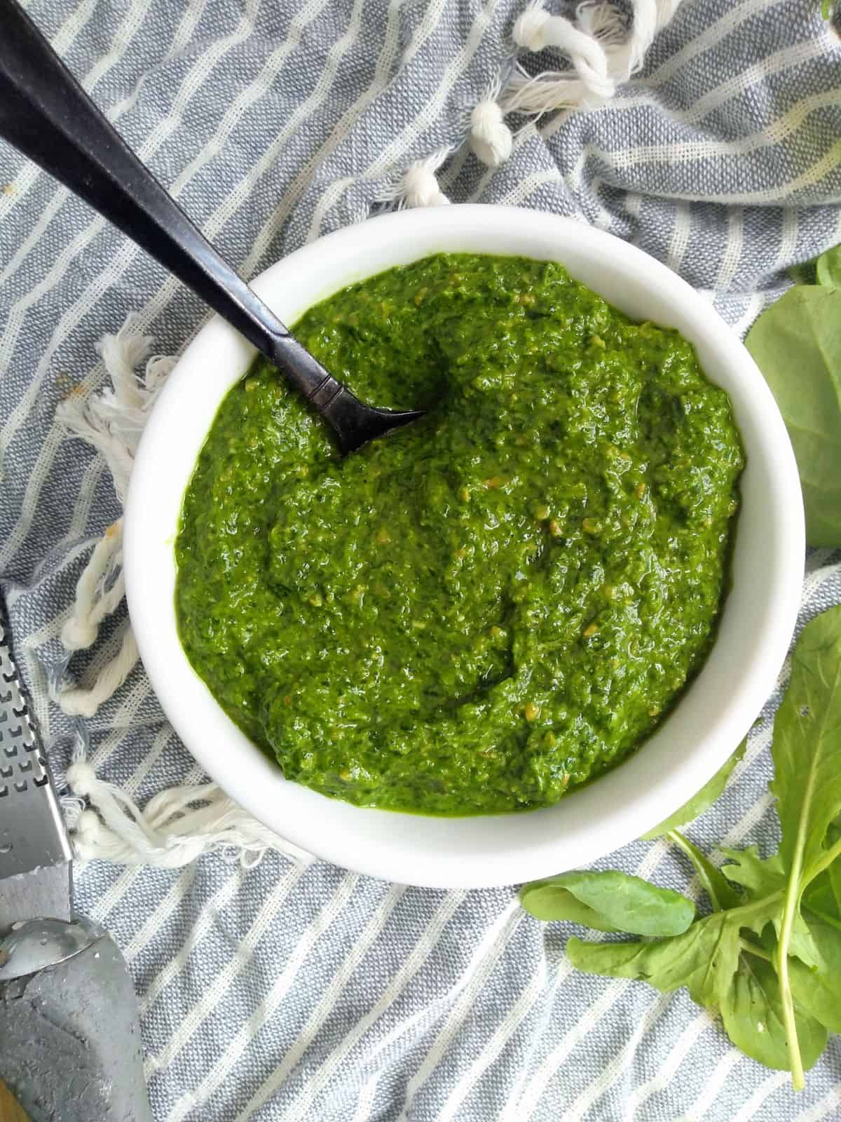 Arugula and Spinach Pesto in a white bowl with a black spoon sticking out, sitting on top of a gray and white striped towel.