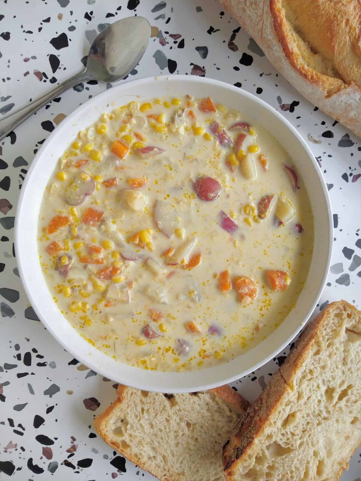 A white bowl filled with corn chowder. Crusty bread and a spoon are next to the bowl.