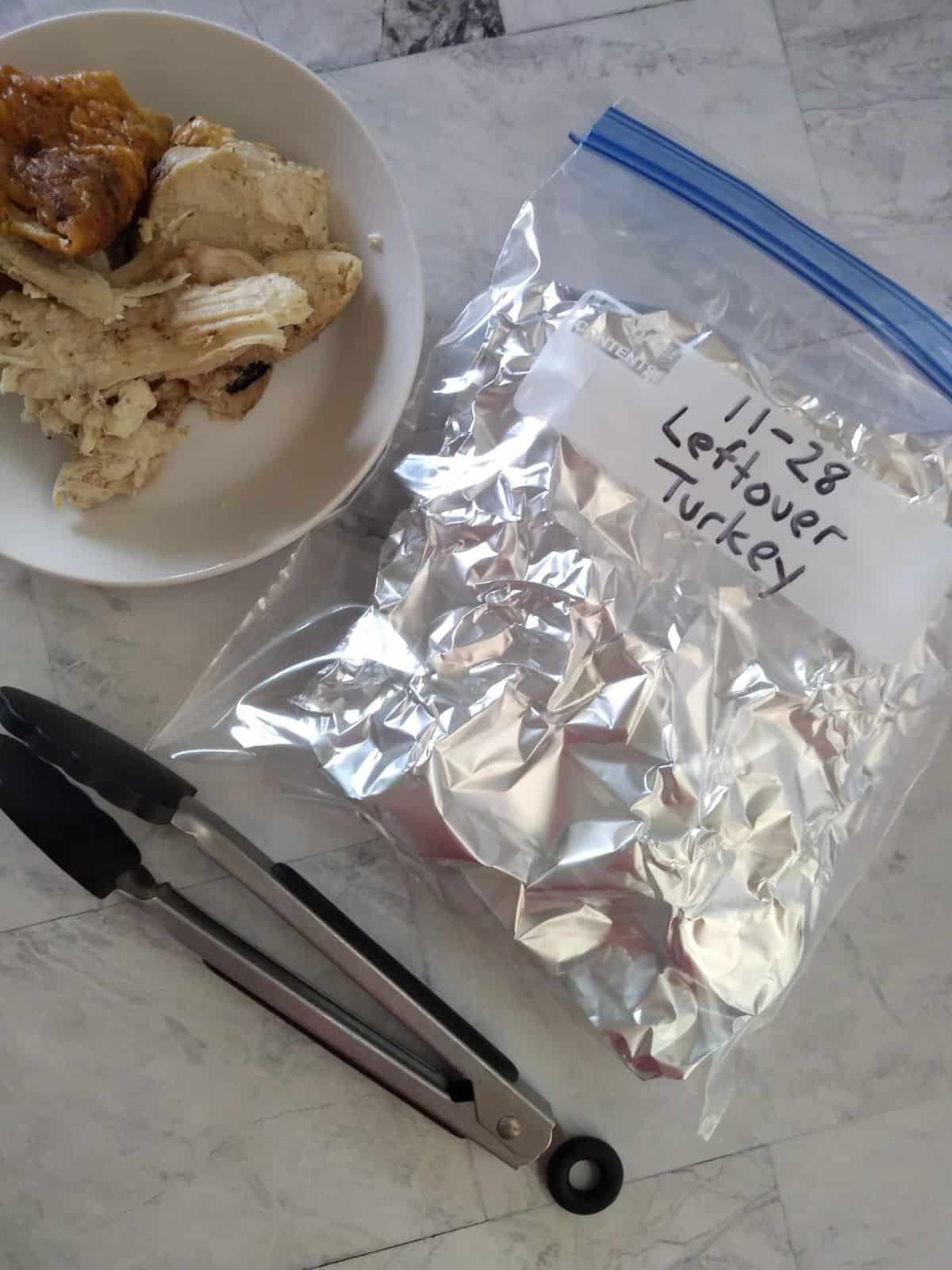 Leftover turkey that has been wrapped in foil that is put inside a plastic bag. A bowl of leftover turkey is next to the bag and a pair of tongs.