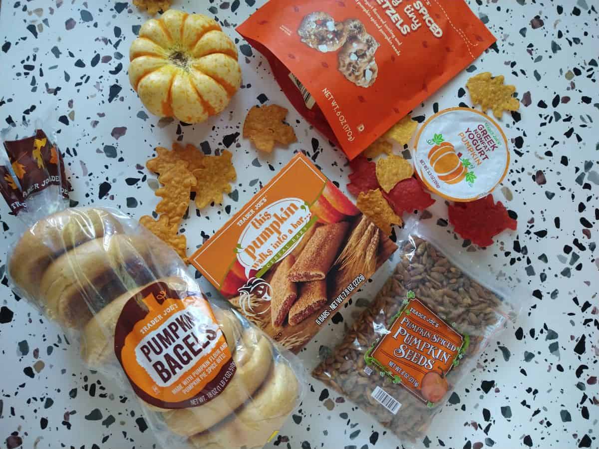 Various Trader Joe's pumpkin products on a table with spots. The products include yogurt, cereal bars, bagels, seeds, and pretzels.