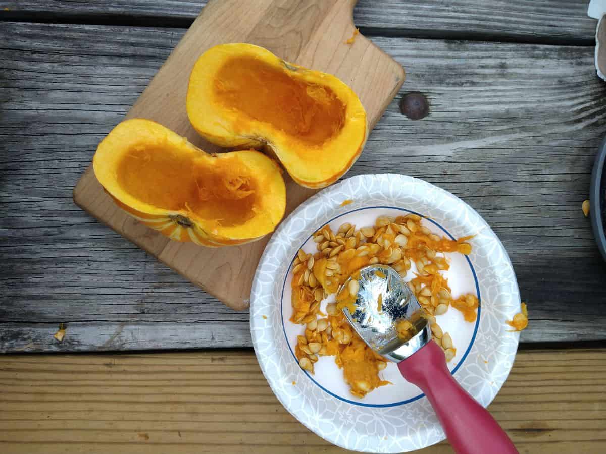 The two halves of the pumpkin on a cutting board with a bowl with the seeds and an ice cream scoop next to the pumpkin. 