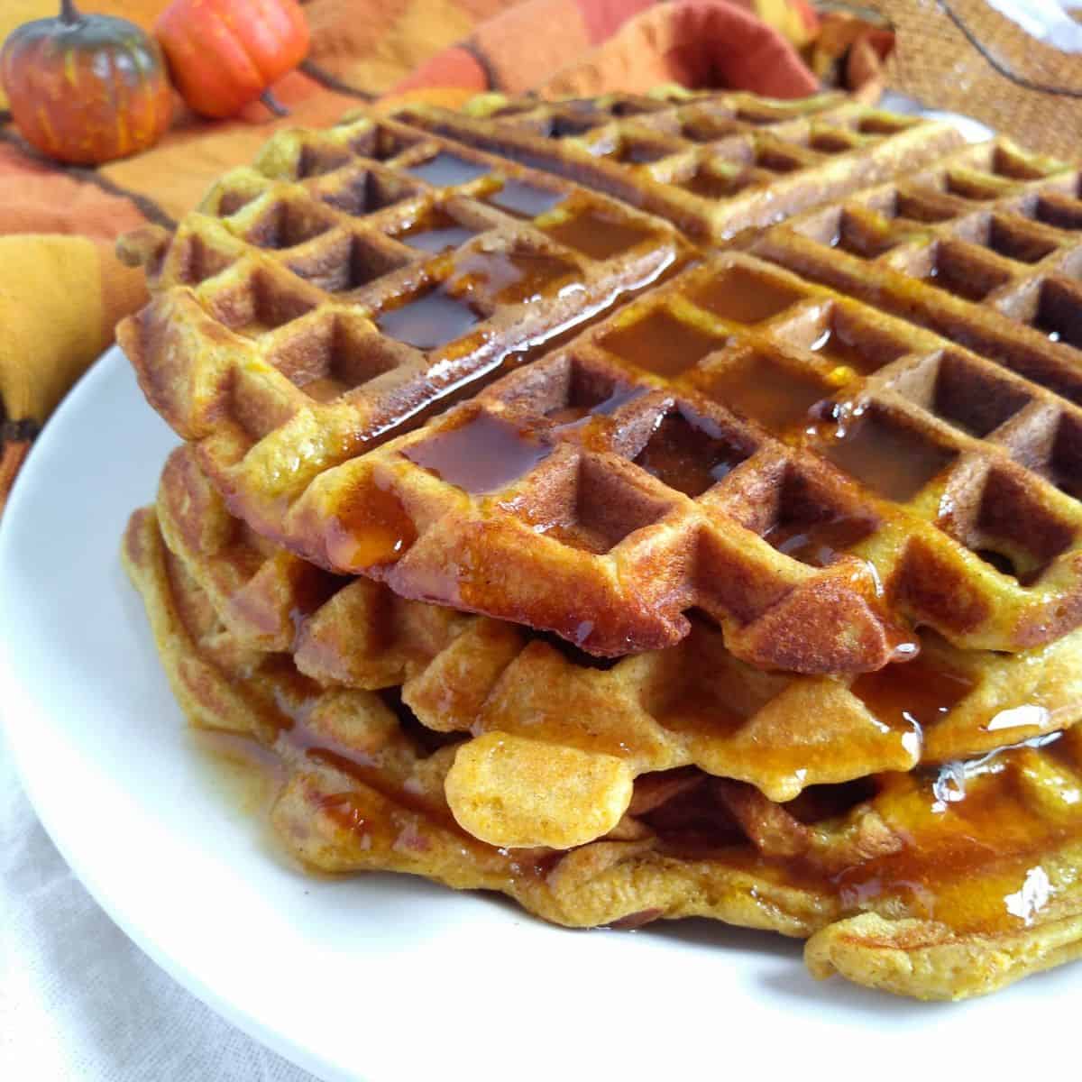 A stack of yeasted pumpkin waffles on a white plate. The waffles are covered in maple syrup.