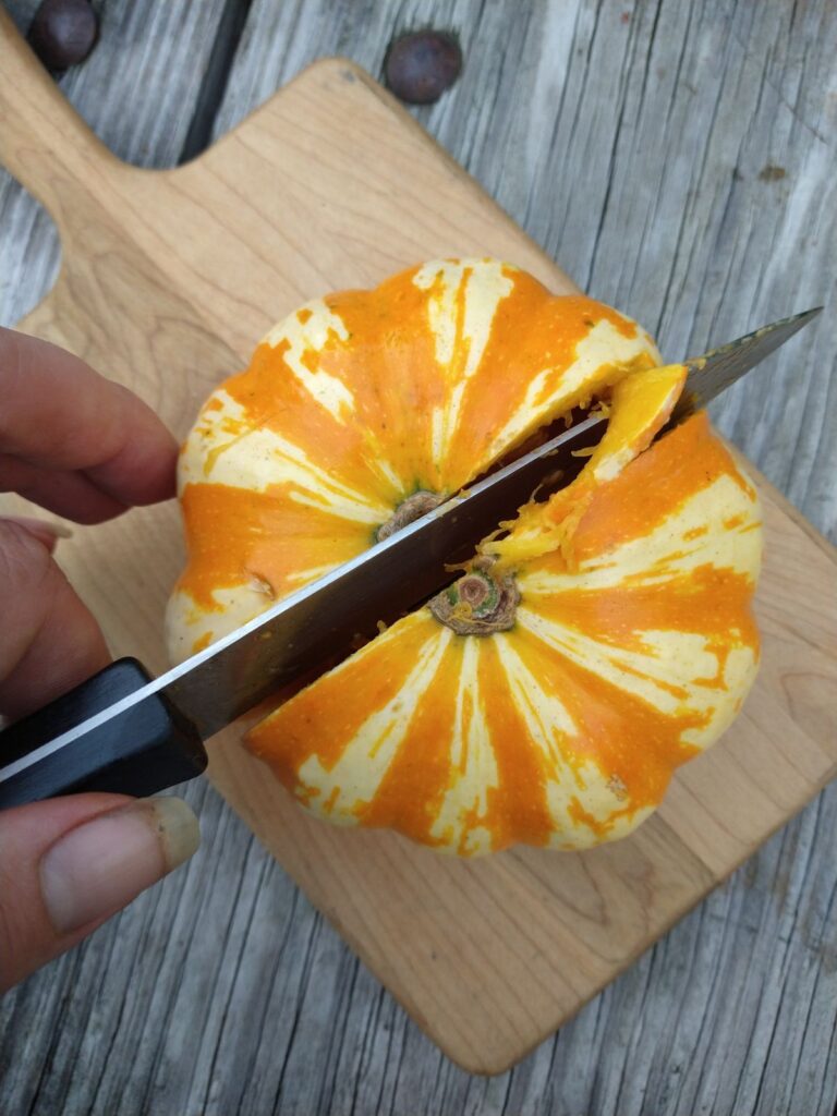 A knife going through the center of the pumpkin. The pumpkin is sitting on a cutting board.