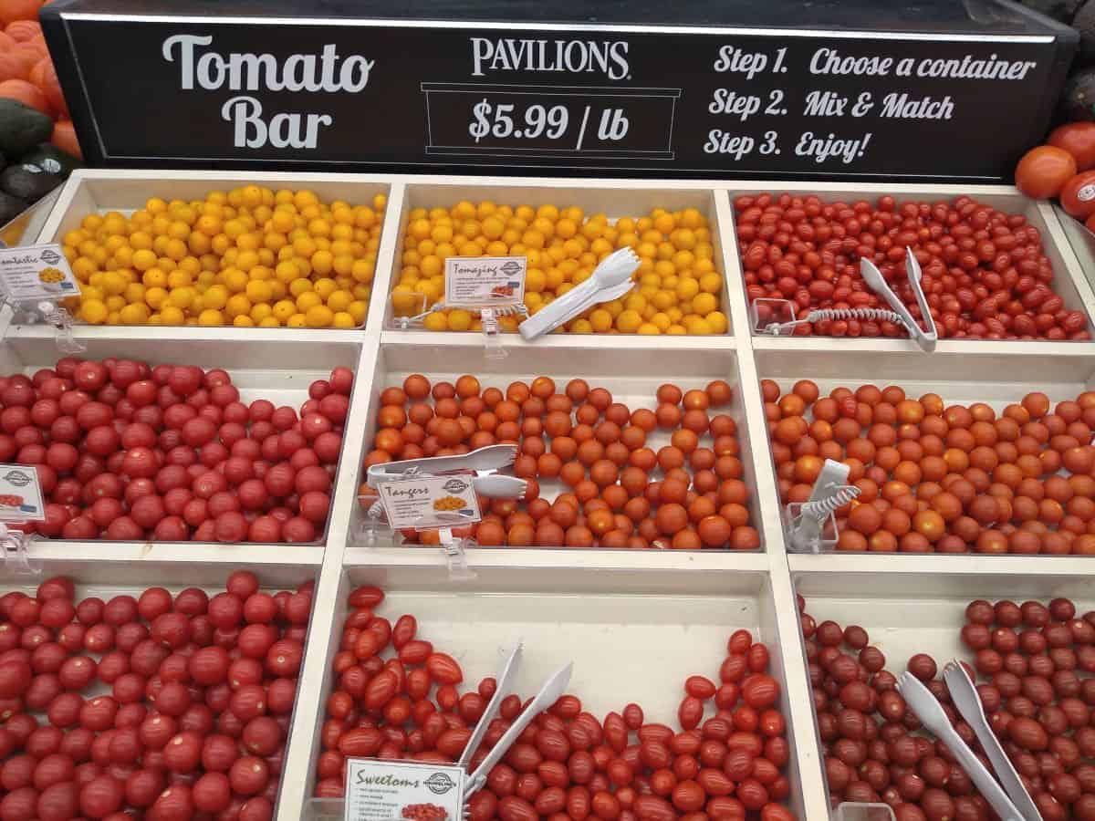 Tomato bar display at a Pavilions grocery store. Tomatoes are all different colors and shapes. A sign marks each variety and a pair of tongs is there to grab them with. 