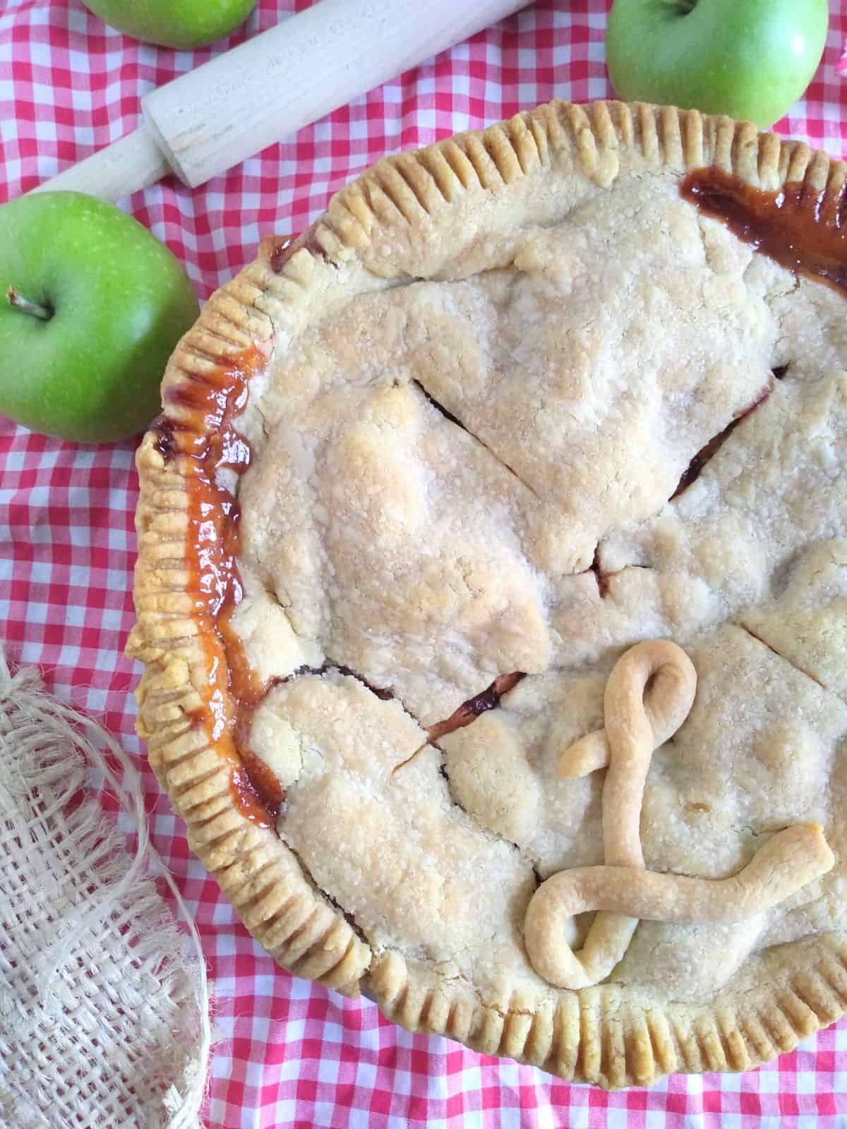 An apple pie with a double crust sitting on a red and white checkered towel. Green apples are next to the pie.