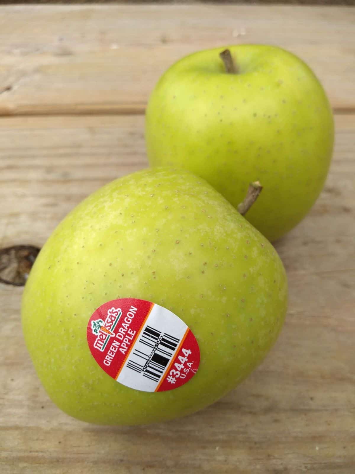 A close up on a table of two Green Dragon apples with a Melissa's PLU sticker on them.