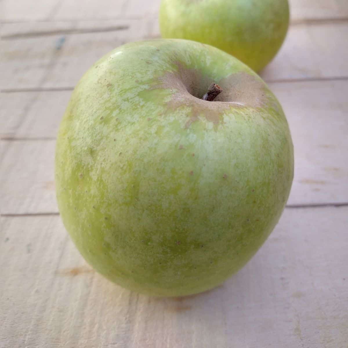 A green Mutsu apple on a wood board with another apple behind it.