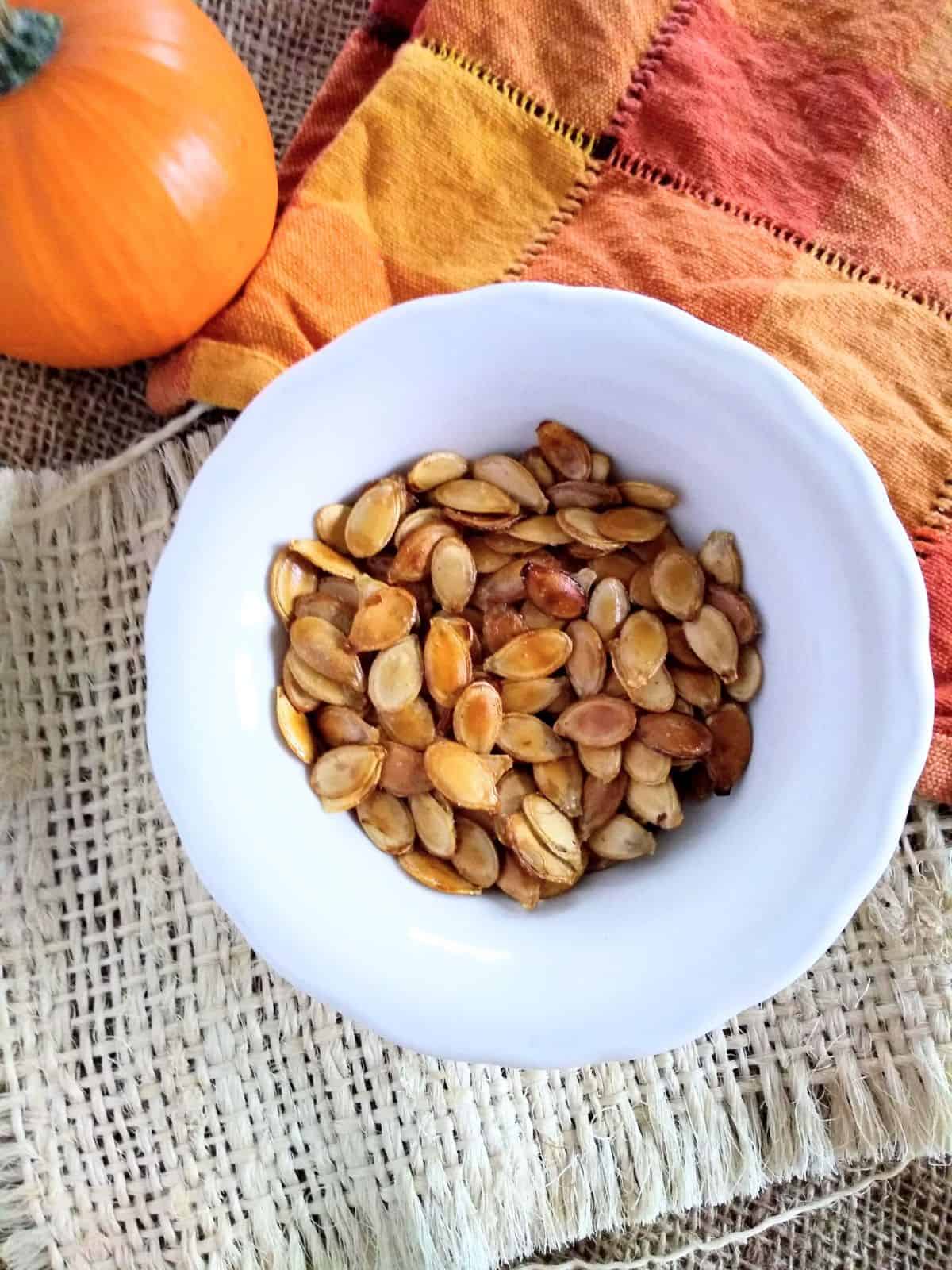 Pie pumpkin seeds that have been roasted sitting in a white bowl with a orange plaid towel underneath.