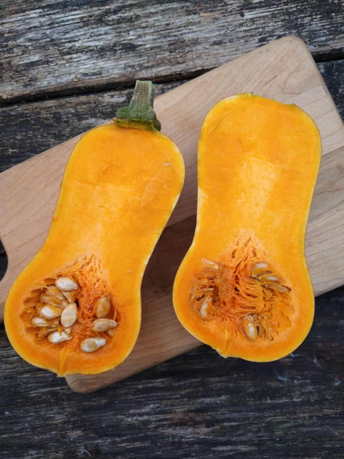 Two halves of a raw Honeynut squash on a wood cutting board on a wood picnic table.