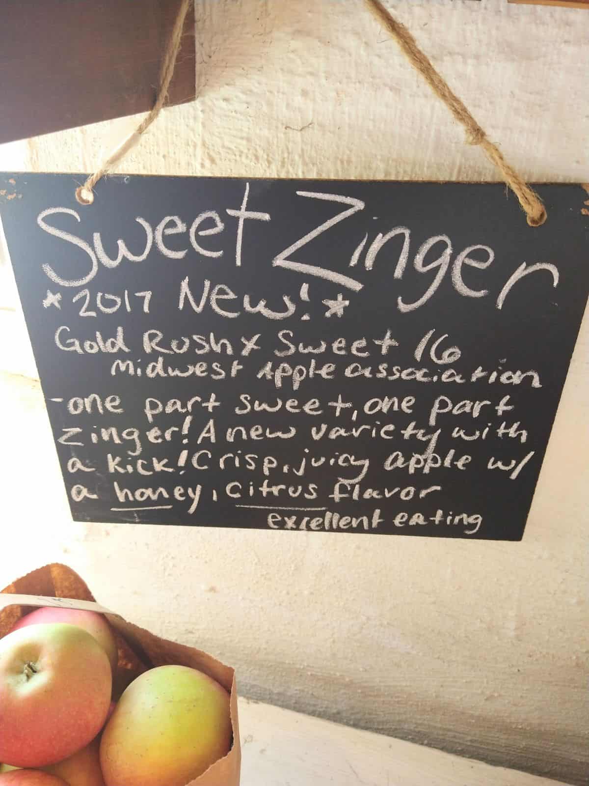 A chalkboard sign that reads "Sweet Zinger - 2017 New! Gold Rush x Sweet 16 Midwest Apple Association. - One part sweet, one part Zinger! A new variety with a kick! Crispy, juicy apple with a honey, citrus flavor. Excellent eating. 