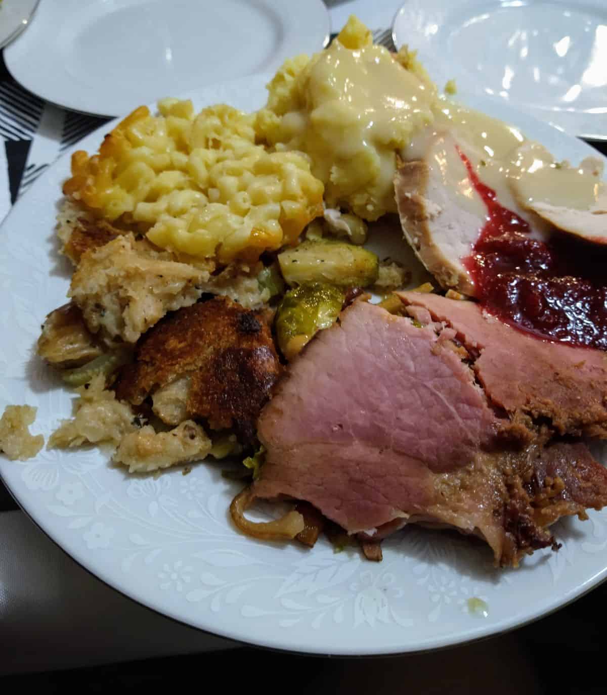 A white plate with Thanksgiving food on it including ham, turkey, cranberry sauce, stuffing, mashed potatoes with gravy, and mac & cheese.