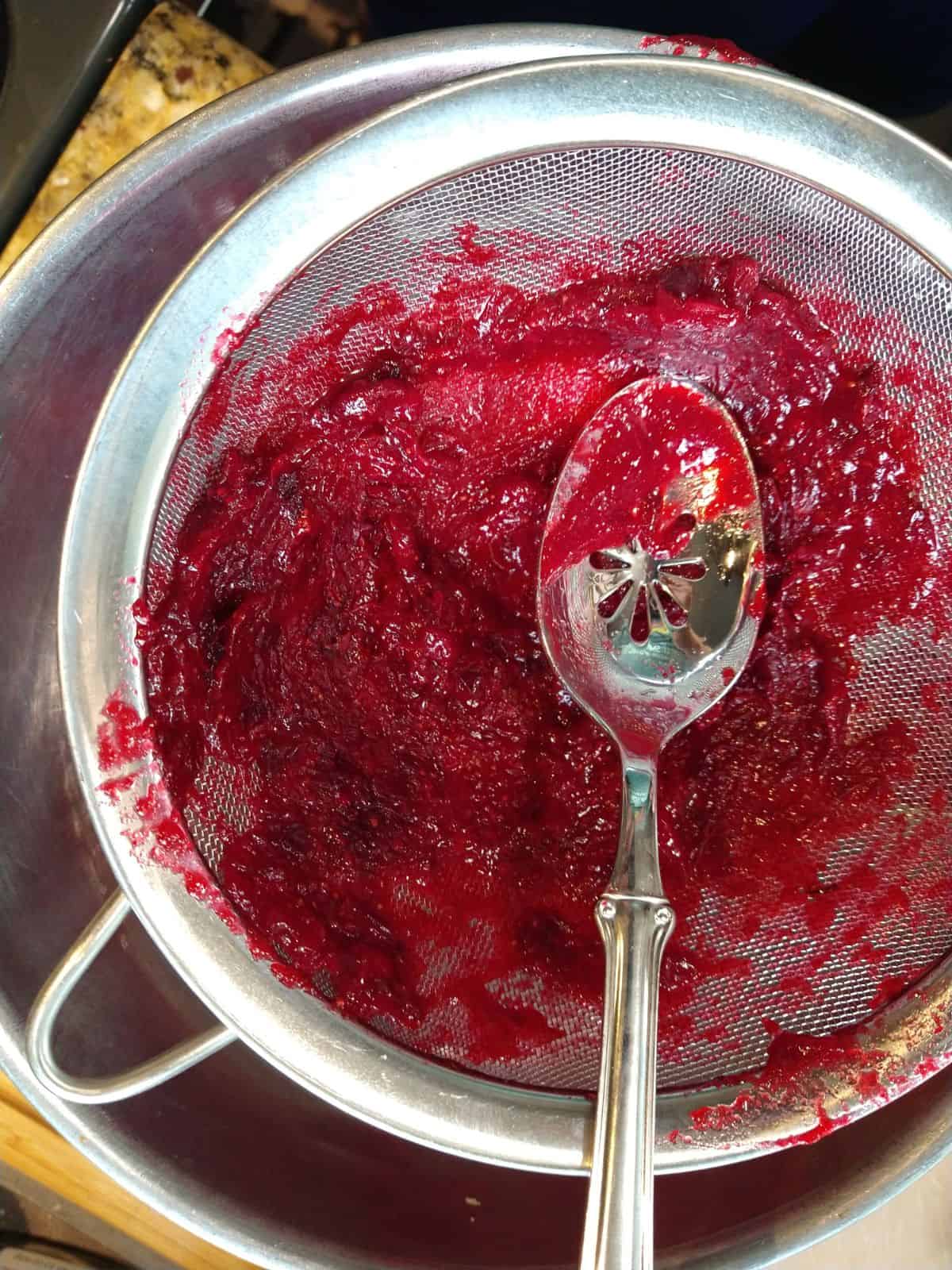A spoon pushing hot cranberry sauce through a sieve into a mixing bowl.