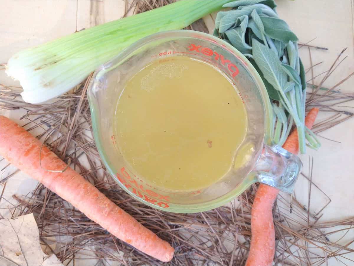 A Pyrex glass measuring cup filled with smoked turkey stock surrounded by celery, sage, carrots, leaves, and pine needles.