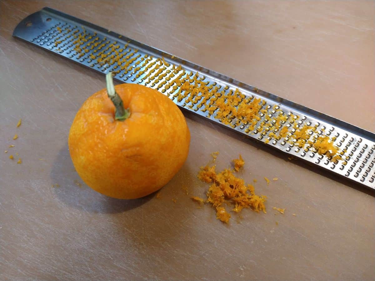 A mandarin with zest removed from it and the zest is on the plastic cutting board and Microplane zester.