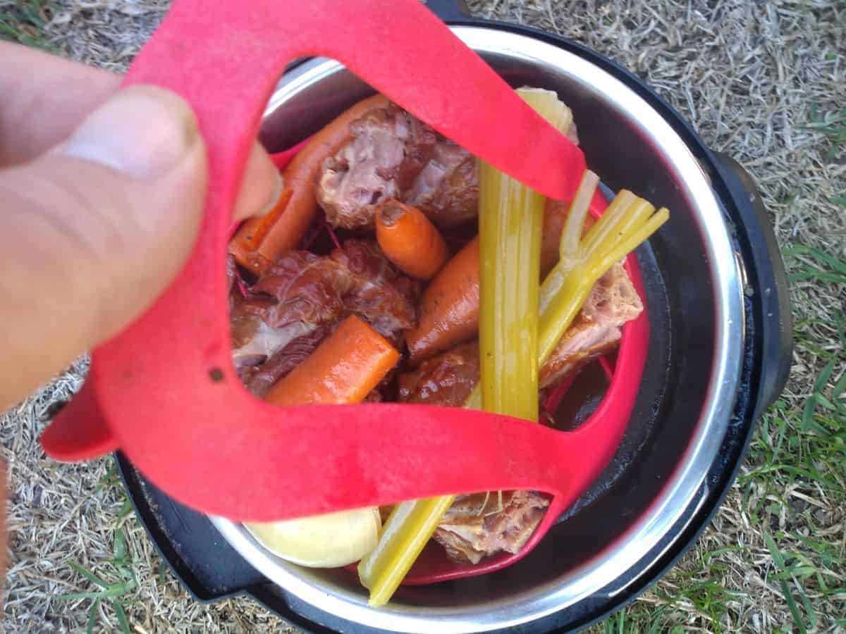 Smoked turkey necks, onion, carrots, celery and thyme are being pulled out of the Instant Pot with a silicone sling.