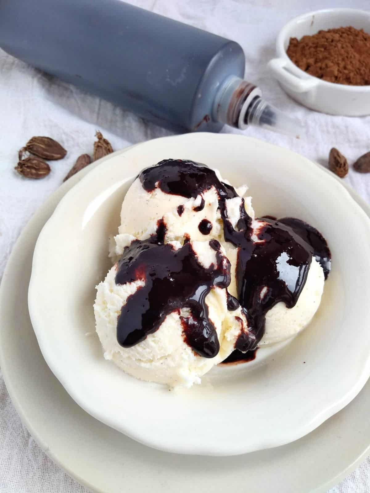 A white bowl with 3 scoop of vanilla ice cream topped with chocolate cardamom syrup. The bottle of syrup is next to the bowl along with whole cardamom pods.