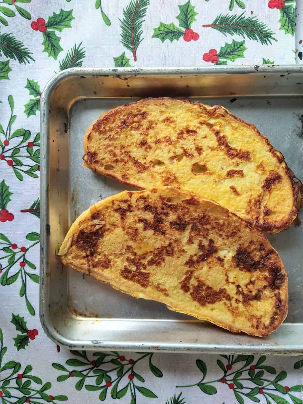 Slices of sourdough French toast on a sheet pan on top of a white cloth with holly on it.