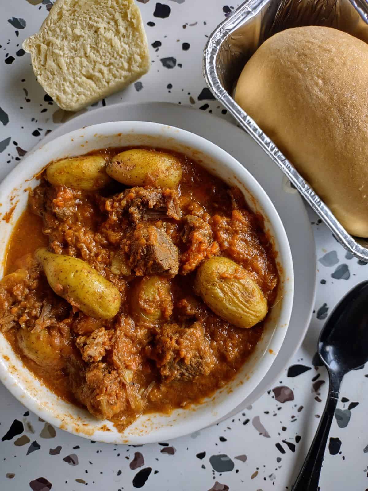 A bowl of beef stew with potatoes and carrots on a white table with dots. A black spoon and a mini loaf of bread sit next to the bowl.