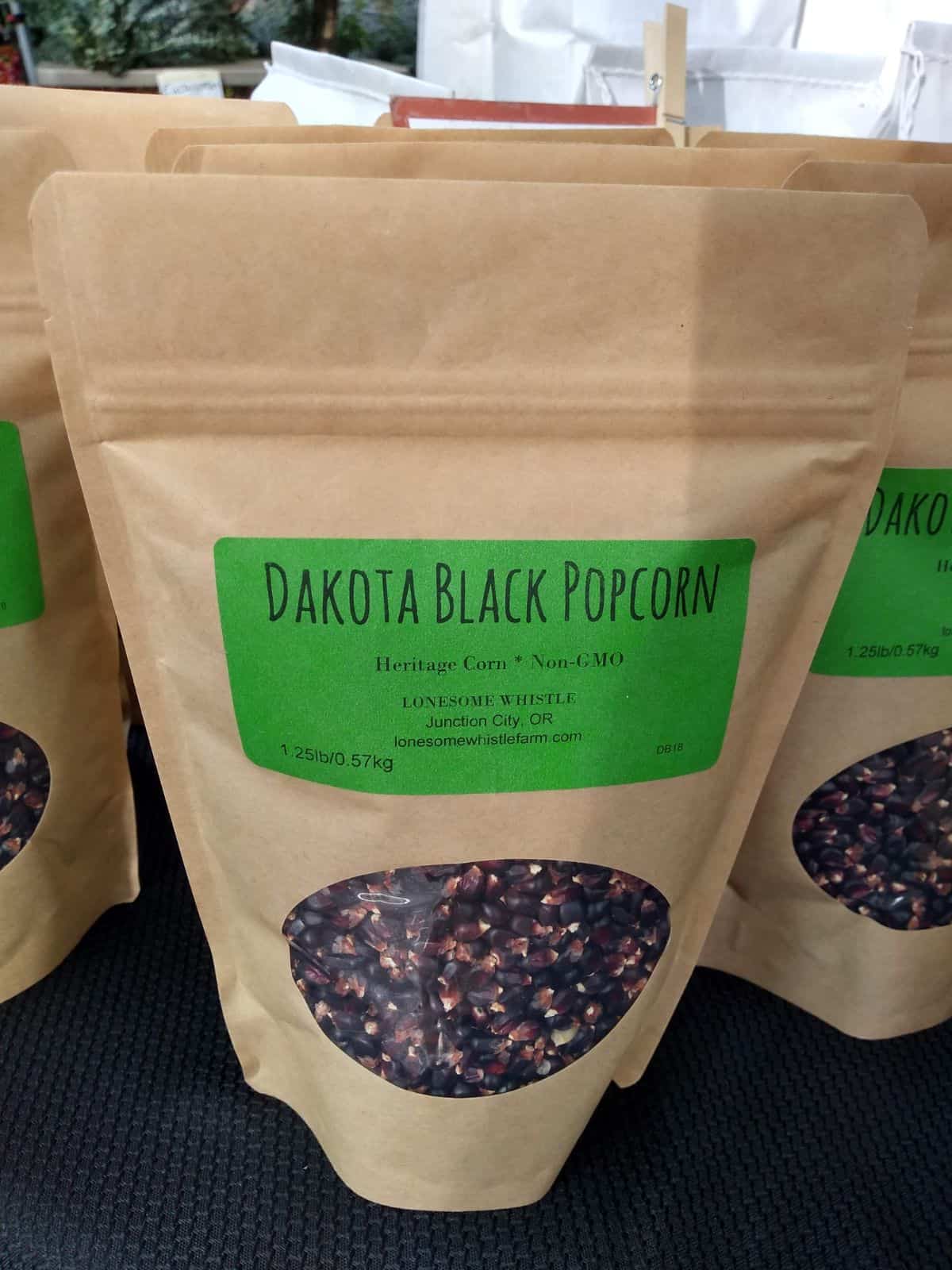 A paper bag with a green label that says Dakota Black Popcorn. You can see through a window the black kernels inside the bag.