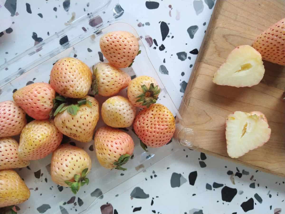 An opened package of white pineberry strawberries next to a cutting board with them sliced open.