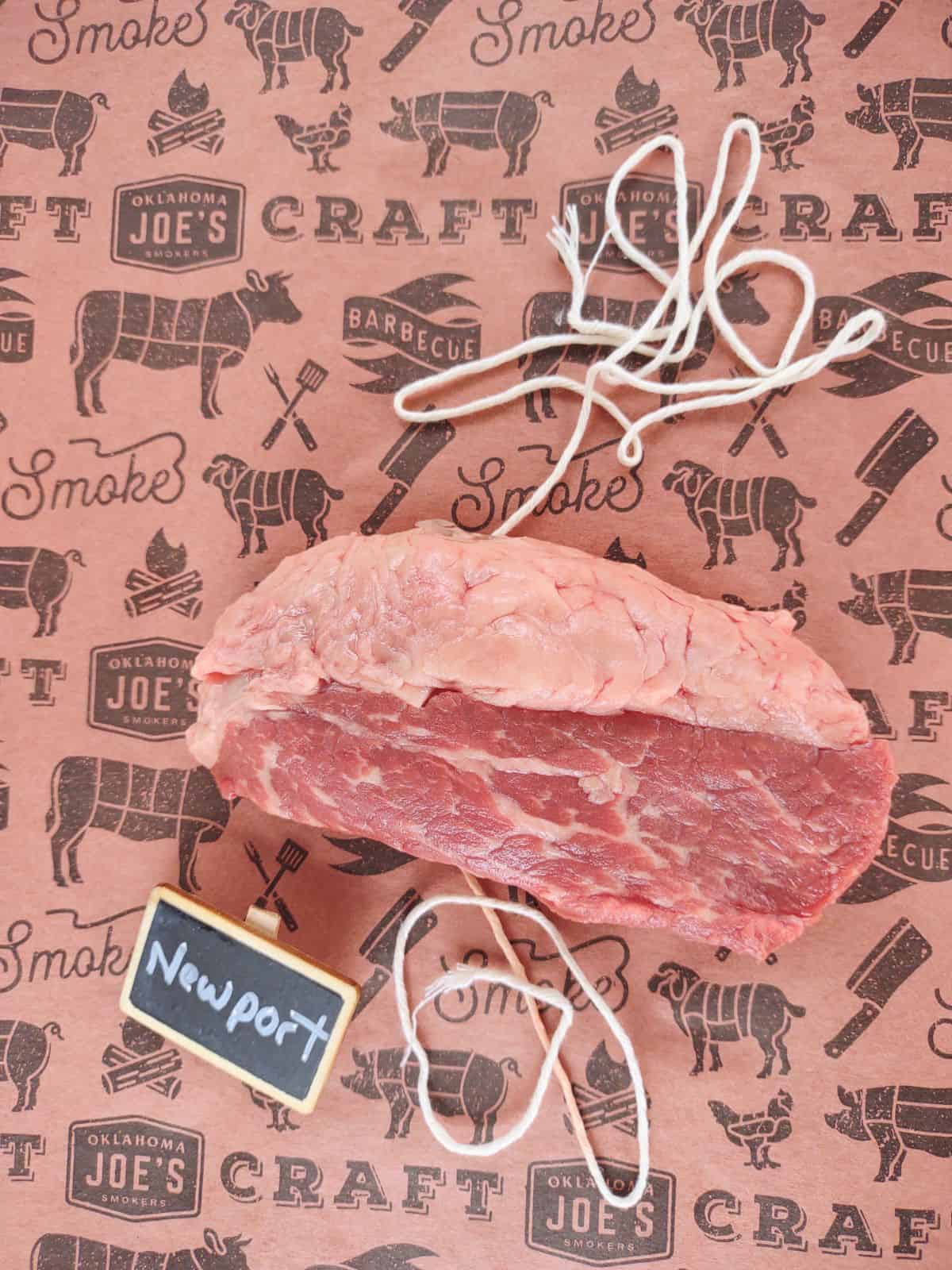 A raw Newport steak on a piece of butcher paper with string underneath the steak. A small chalkboard sign next to it reads Newport.