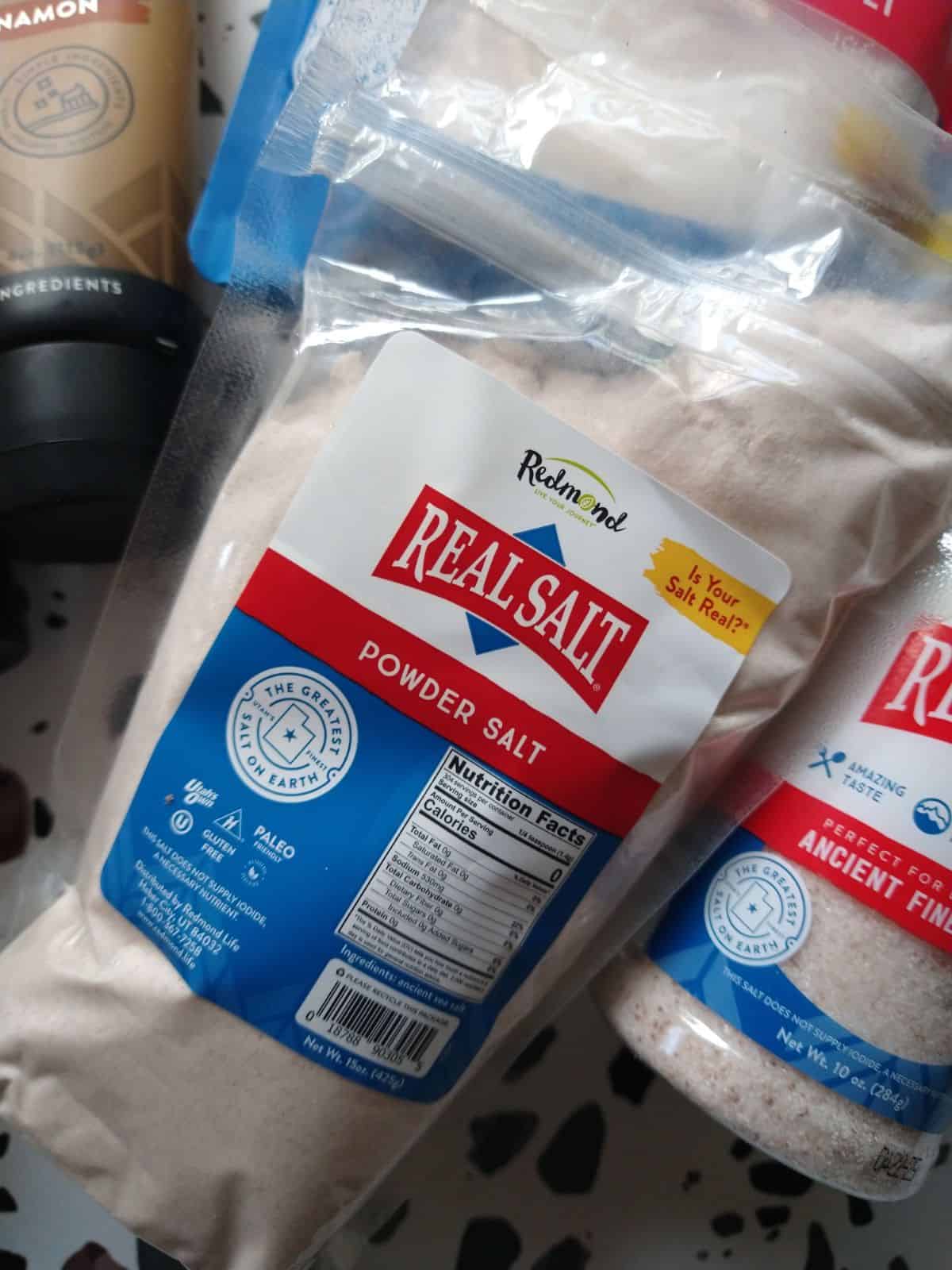 Bags of Redmond Real Salt Powder Salt and a container of fine Sea salt.