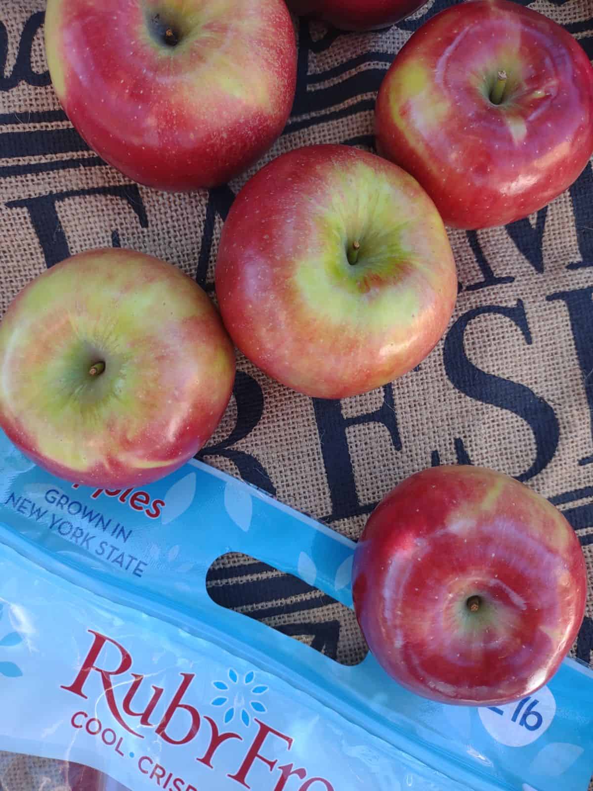 FIve RubyFrost apples on top of a piece of burlap with writing on it. The bag they came in is shown in the bottom left corner.