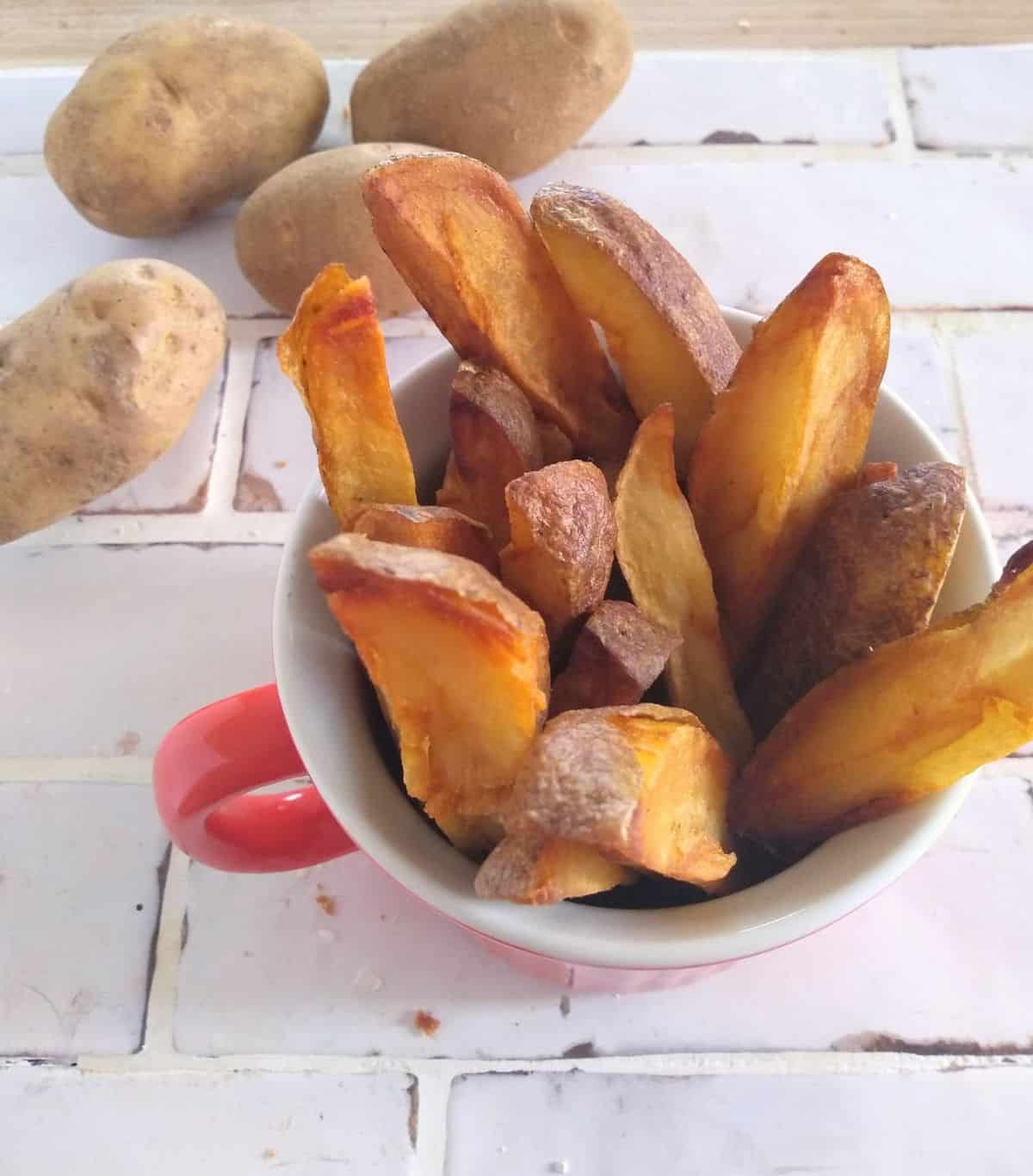 Baked potato fries in a red mug sitting on a white tile countertop with Russet potatoes in the top left corner.