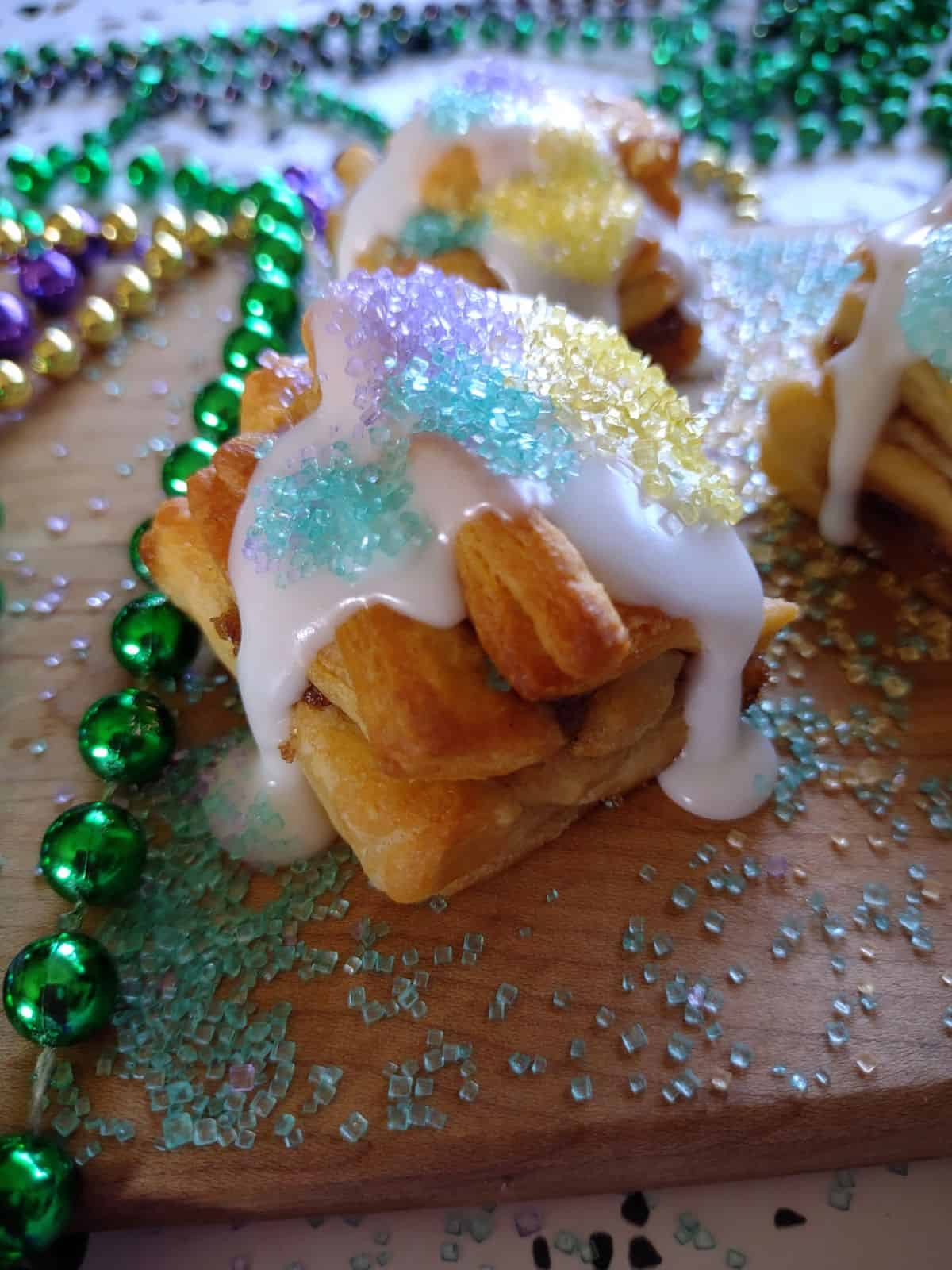 A side view of the layers of mini king cakes made from Crescent roll dough. It's topped with icing with green, yellow. and purple sanding sugars. Mardi Gras beads are in the background.