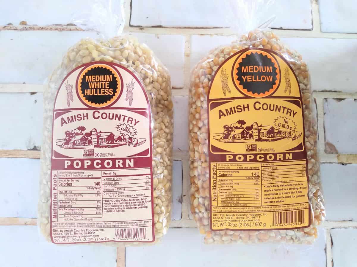 Bags of Amish Country bagged popcorn - Medium White Hulless and Medium Yellow, sitting on a white tile countertop.