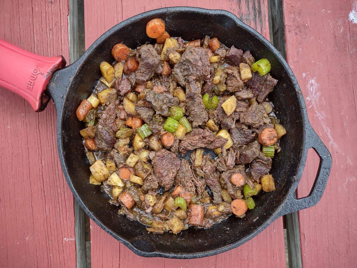 A beef stew filled cast iron skillet with a red handle is sitting on a red picnic table. 