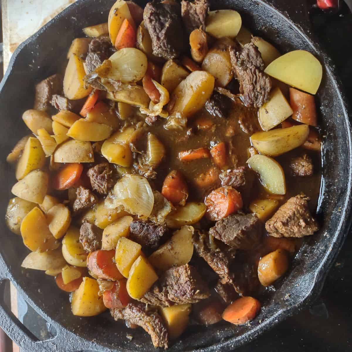 A cast iron skillet filled with beef stew eat, potatoes, parsnips, and carrots. You can see liquid in the bottom of the skillet.
