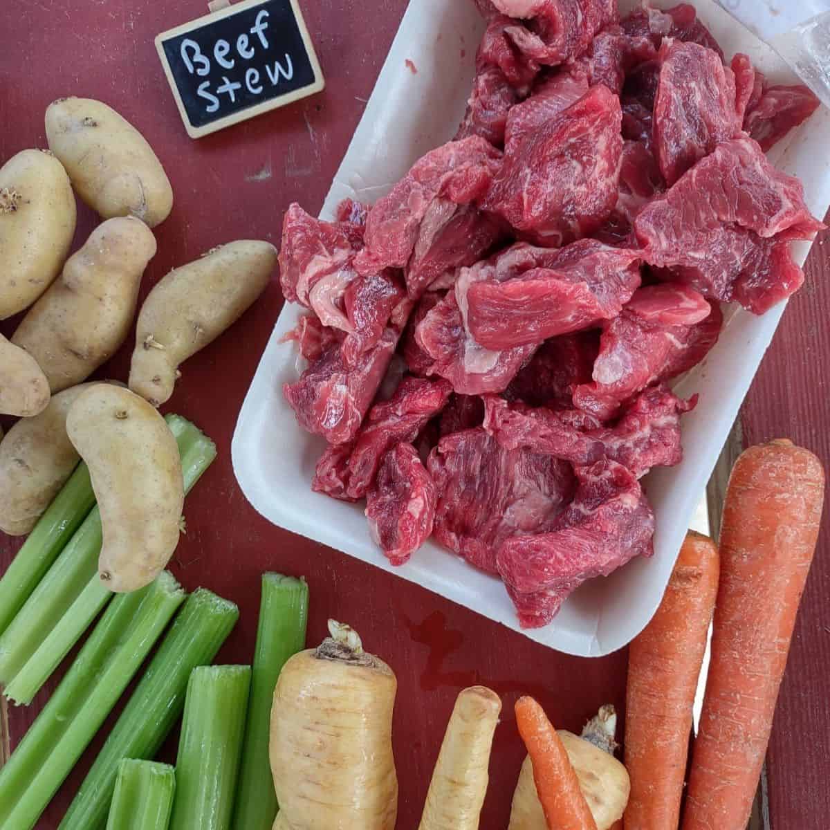 Raw beef stew meat with carrots, parsnips, celery, and Fingerling potatoes on a red table.