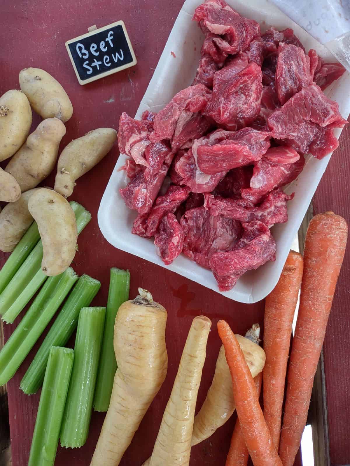 Raw beef stew meat, fingerling potatoes, celery sticks, parsnips, and carrots are arranged on a red picnic table. 
