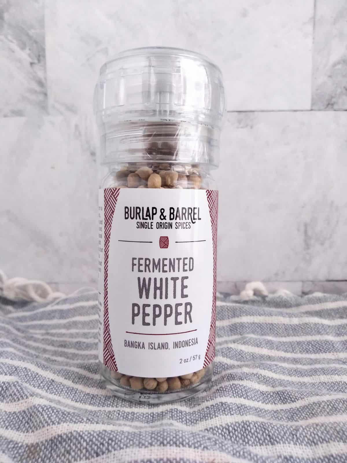 Fermented White Peppercorns in a jar with a grinder in front of a white tile board.