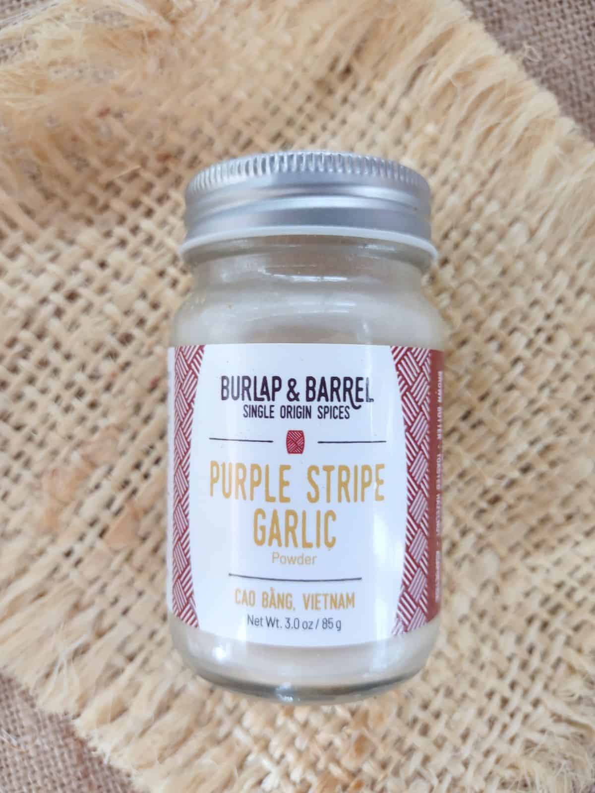 Burlap & Barrel Purple Stripe Garlic in a glass jar sitting on a piece of burlap on top of a darker piece of burlap.
