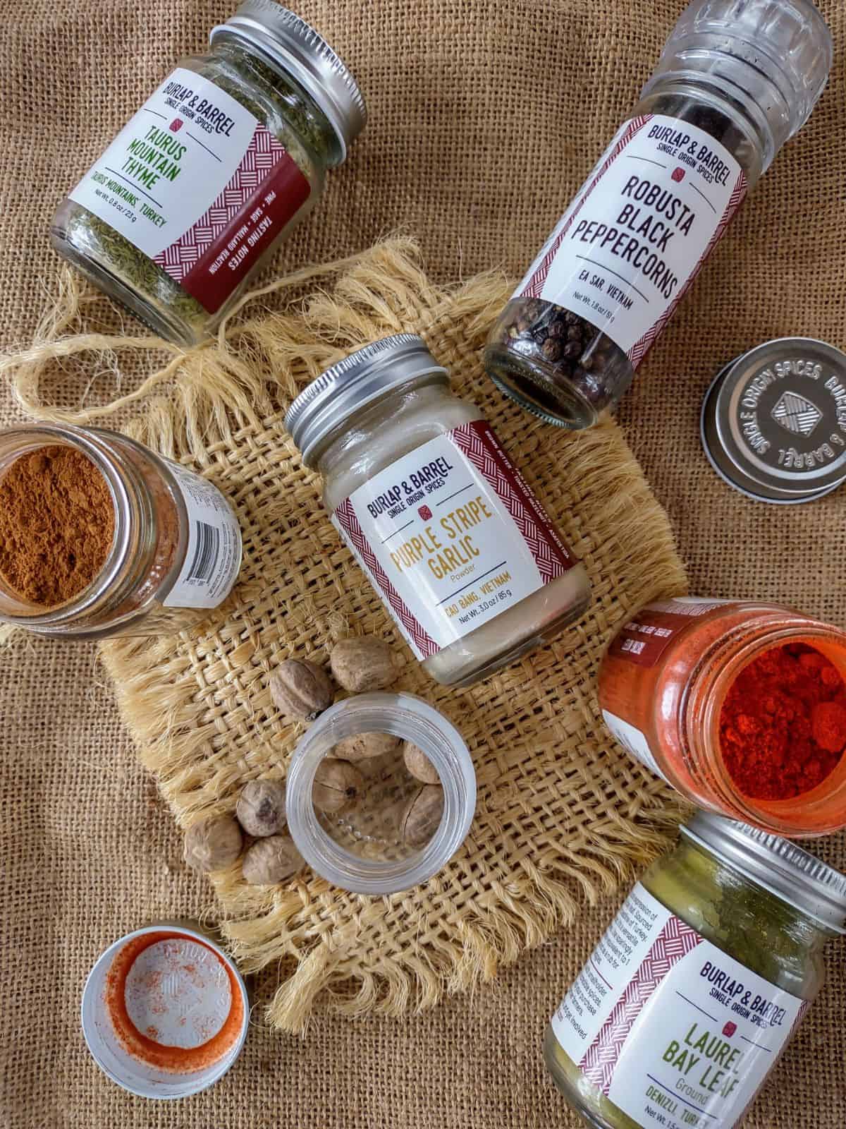 Various Burlap and Barrel Spices in jars and grinder siting on two different pieces of burlap.