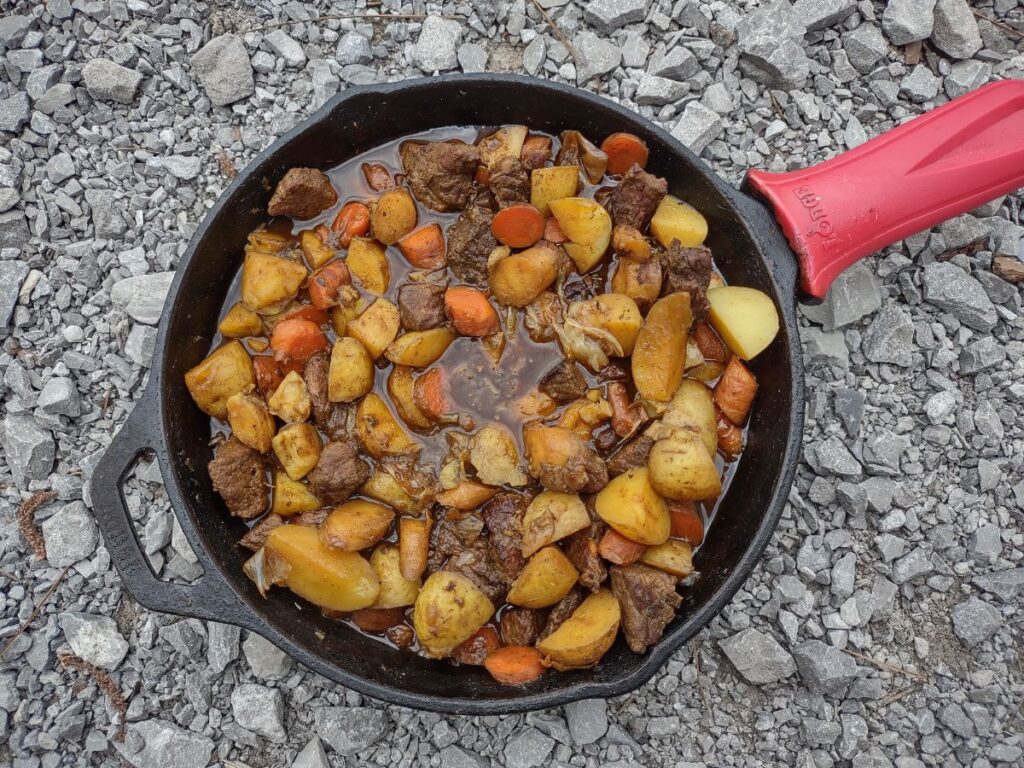 Beef stew with carrots, potatoes, parsnips, and pearl onions in a cast iron skillet with a red handle sitting on gray gravel.