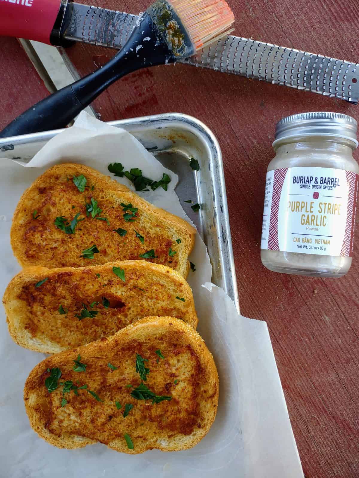 A sheet pan with 3 slices of garlic bread stacks on each other. The bread is topped with parsley. There is a container of Burlap & Barrel Purple Stripe Garlic powder to the right of the pan.