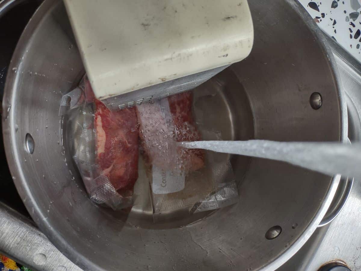 A white immersion circulator attached to a stock pot with two steaks wedged underneath the circulator. Water is being added to the pot.