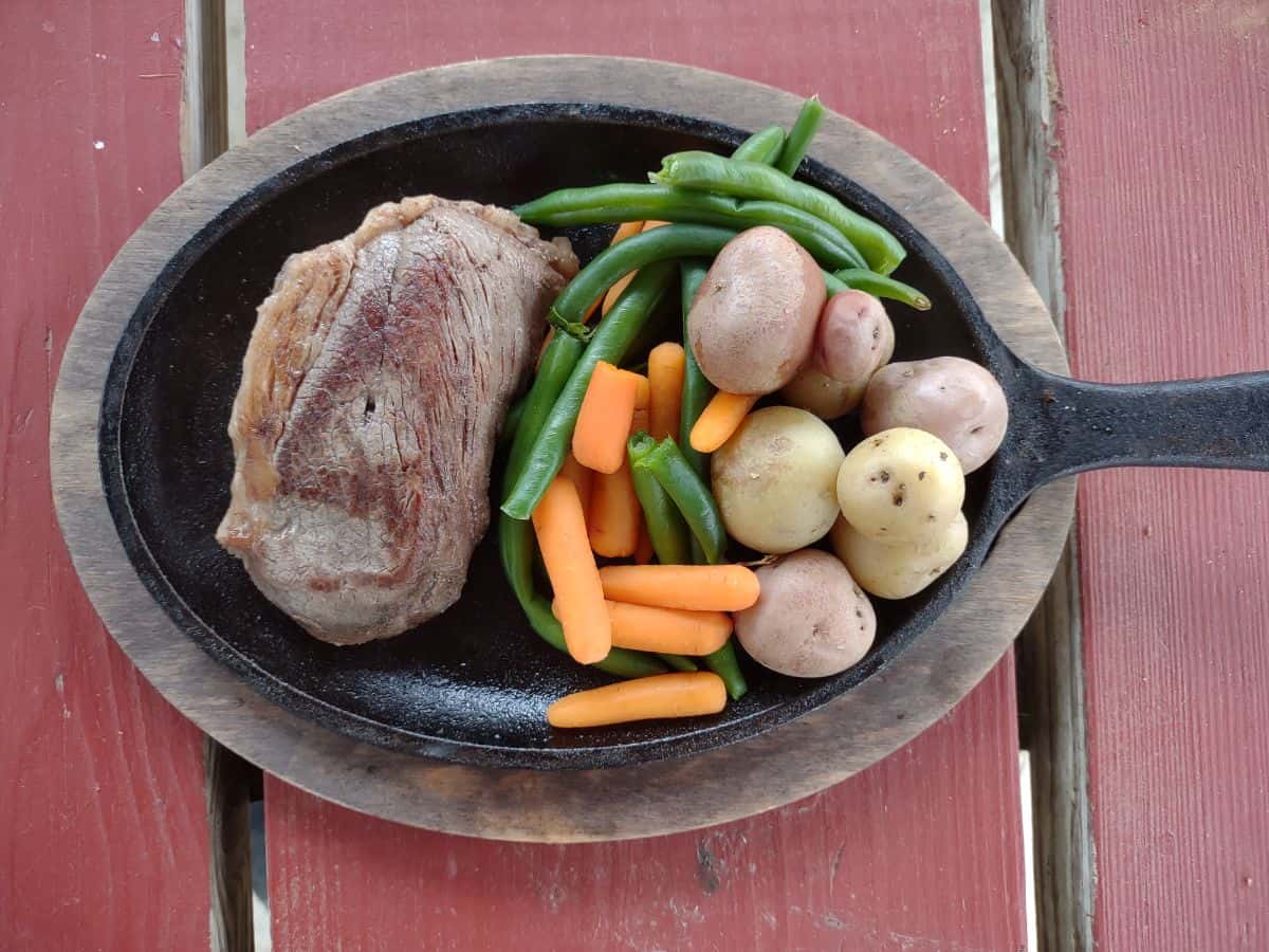 A cast iron griddle with a steak, small whole potatoes, green beans, and baby carrots.