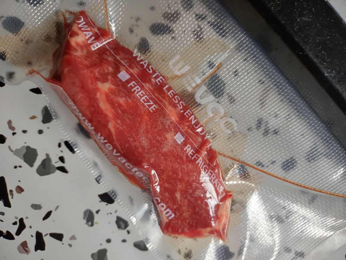 A close up of a vacuum sealed Petite Denver steak sitting on a white tale with spots. 