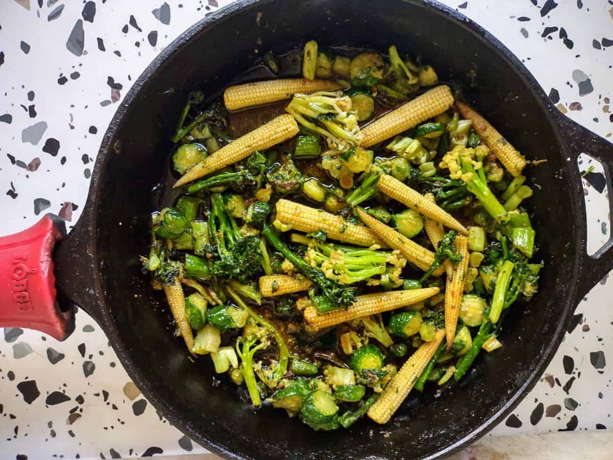 A black cast iron skillet with a red handle filled with a stir fry of baby corn, baby zucchini, baby broccoli, and baby cauliflower.
