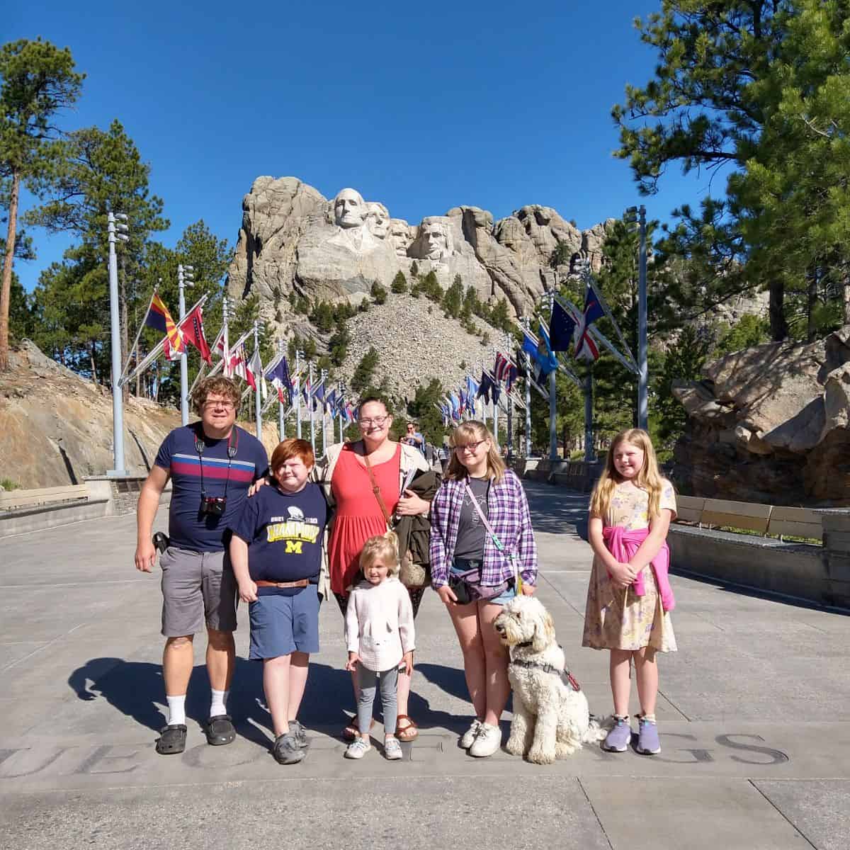 A family of 6 and a dog standing in front of Mt Rushmore.